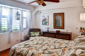 Un dormitorio con cama, ventilador de techo, ventana con cortinas, una silla de mimbre, cómoda, espejo y un cuadro.