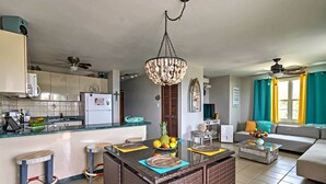 A modern kitchen with a central island, a dining area with bar stools, a living room with a sofa and coffee table, and a ceiling fan.