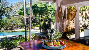 Un comedor con una mesa dispuesta para dos personas, un amplio ventanal con vistas a una piscina y abundante vegetación, y un arreglo decorativo de plantas sobre la mesa.