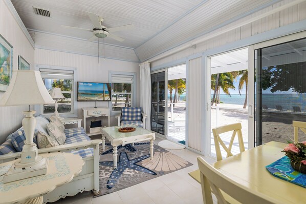 A beachside room with a large sliding glass door to the beach, a ceiling fan, a TV, a dining table, and a sofa with a starfish rug.