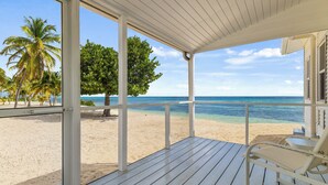 Eine Holzterrasse mit weißer Reling, die auf einen Strand mit Palmen und den Ozean blickt.