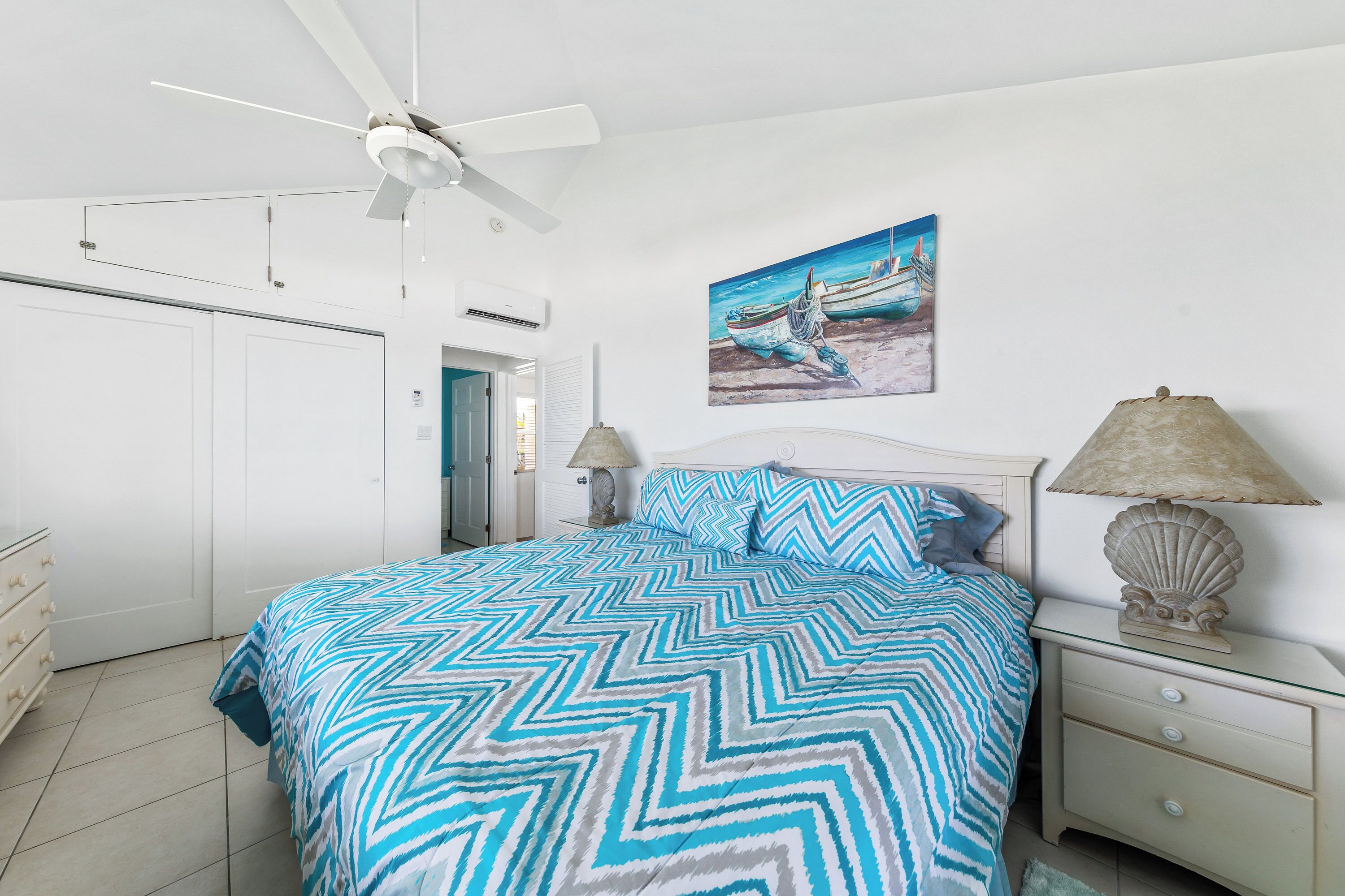 A bedroom with a bed, a ceiling fan, a nightstand, a lamp, and a painting on the wall.
