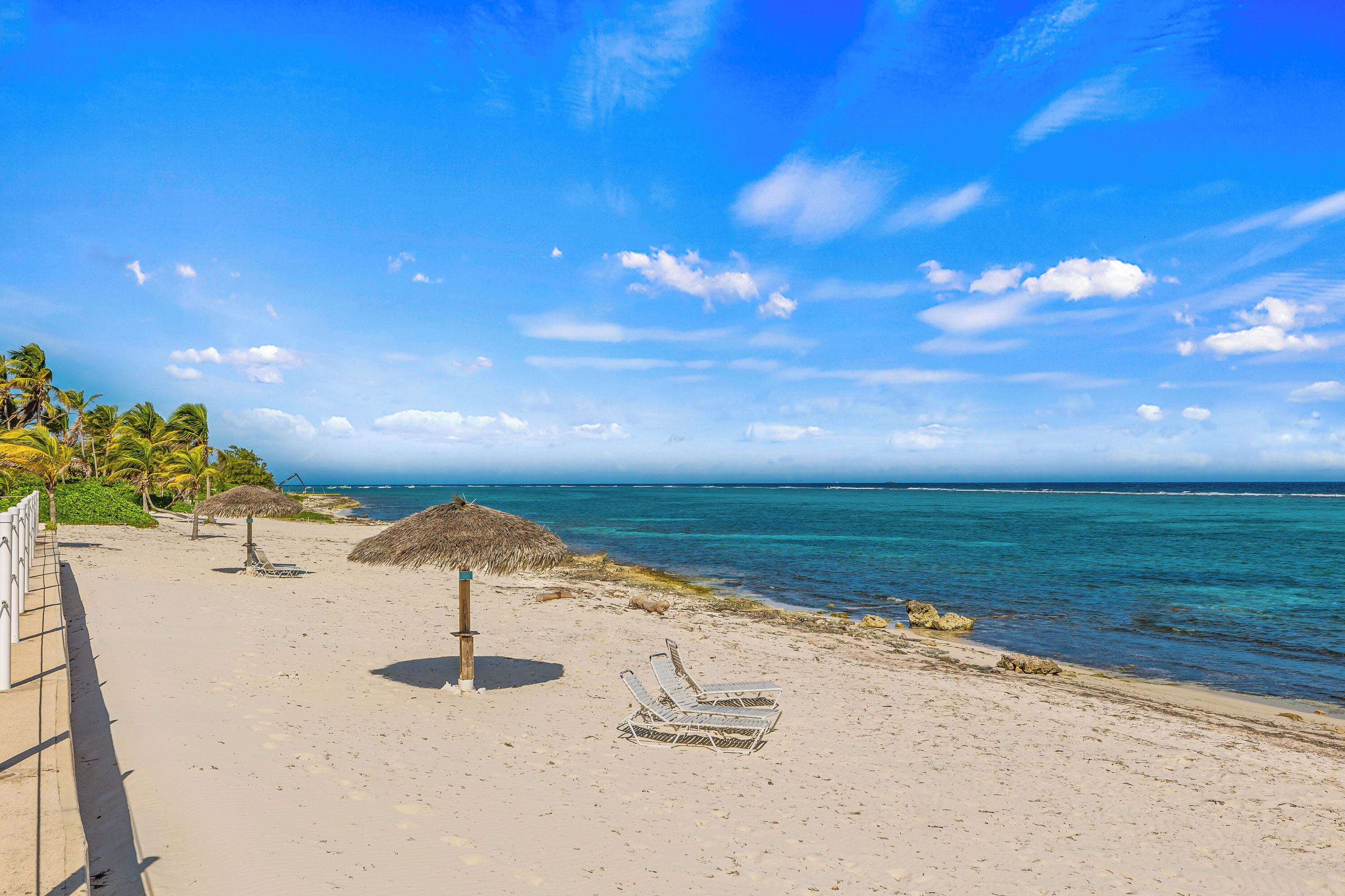 A beach with a thatched umbrella, lounge chairs, and palm trees.