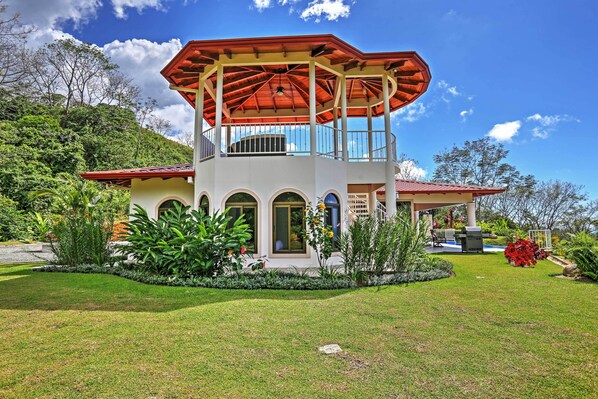A two-story building with a red roof and white walls, surrounded by a well-maintained garden and a hilly landscape.