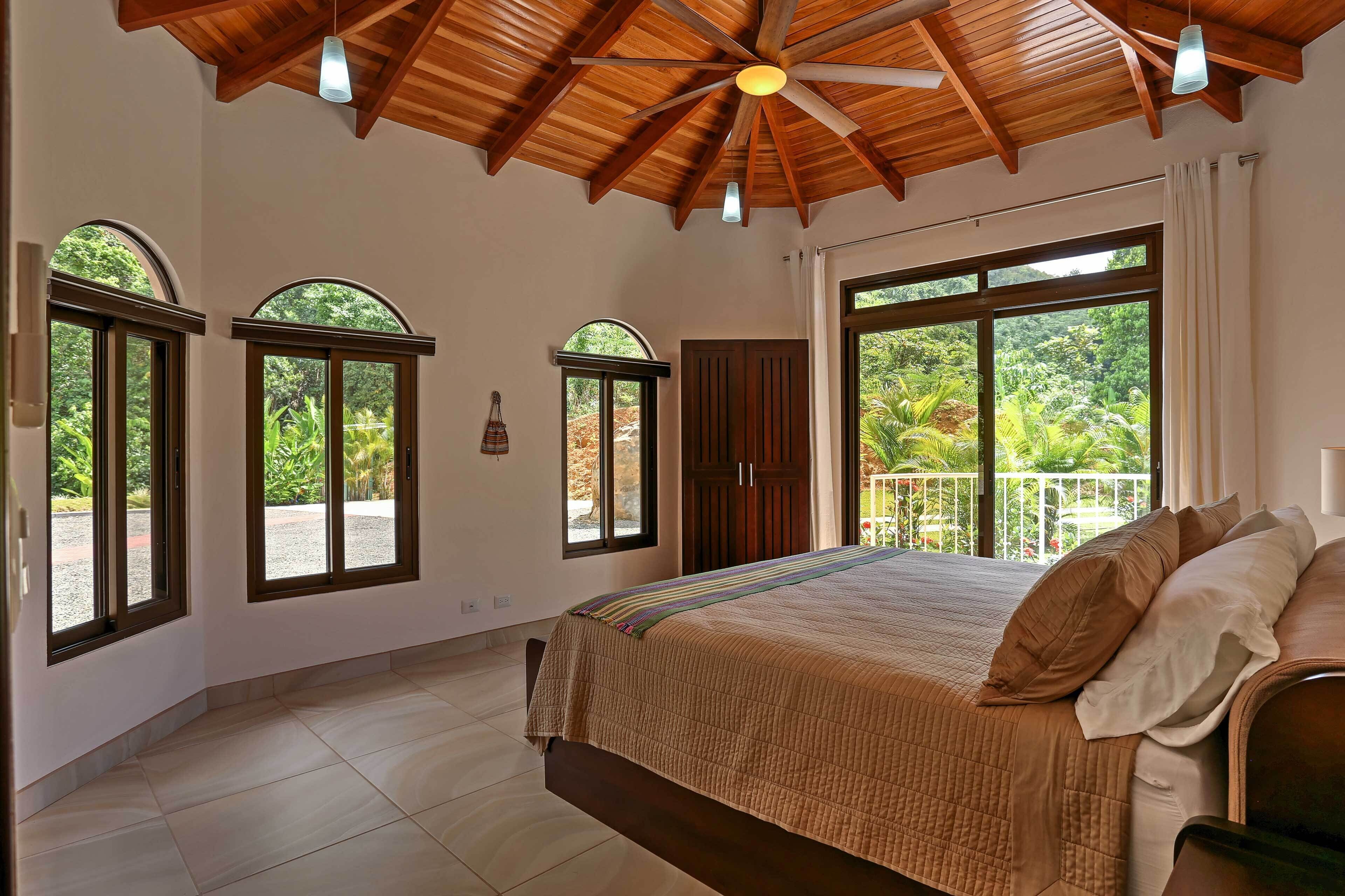 A spacious bedroom with a large bed, wooden ceiling, and large windows offering a view of greenery.