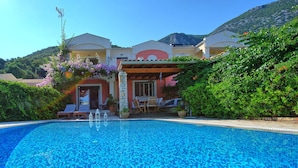 A swimming pool with a clear blue mosaic tile design, surrounded by lush greenery and a building with a covered patio area.