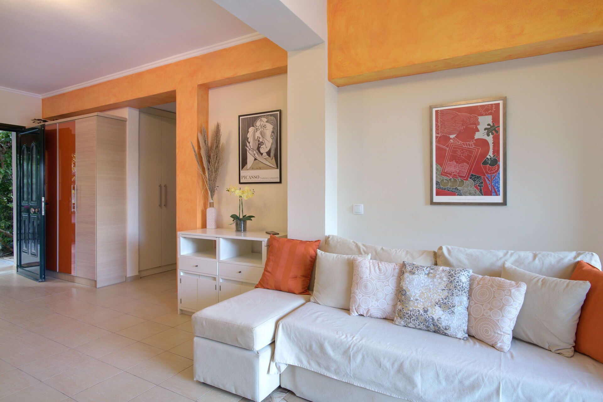 A modern living room with a white sofa, orange cushions, a white coffee table, and a framed Picasso painting.