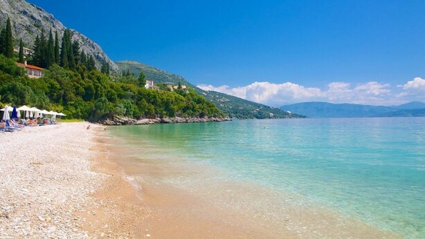 A beach with clear turquoise water, a pebble beach, and a hillside with buildings.