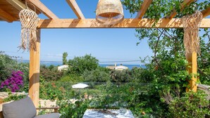 A wooden pergola with woven lampshades, a table with a patterned tablecloth, and a view of the sea and buildings in the background.