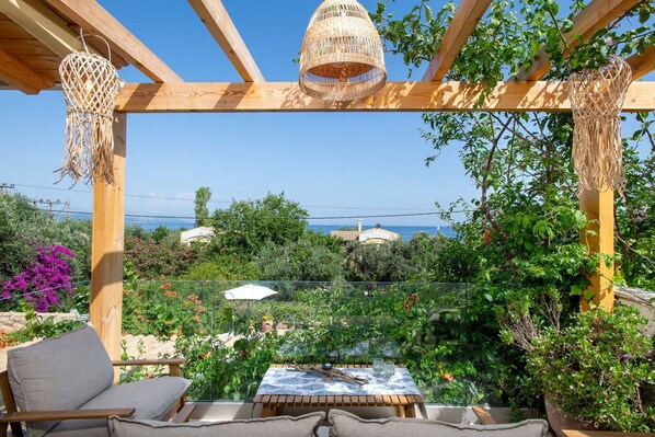 A wooden pergola with woven lampshades, a table with a patterned tablecloth, and a view of the sea and buildings in the background.