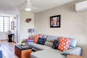 A living room with a grey sofa, colorful cushions, a framed cityscape painting, and a small table with a plant.