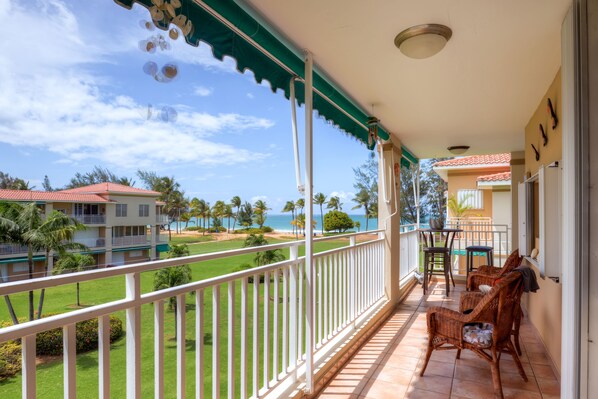 A balcony with wicker chairs, a table, and a view of the ocean and buildings.