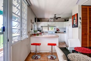 Une cuisine de taille réduite avec des meubles blancs, un îlot central, deux tabourets de bar rouges et un ventilateur de plafond.