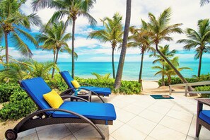 Un salon de plage avec des fauteuils bleus rembourrés et une petite table, entouré de palmiers et offrant une vue sur l’océan.