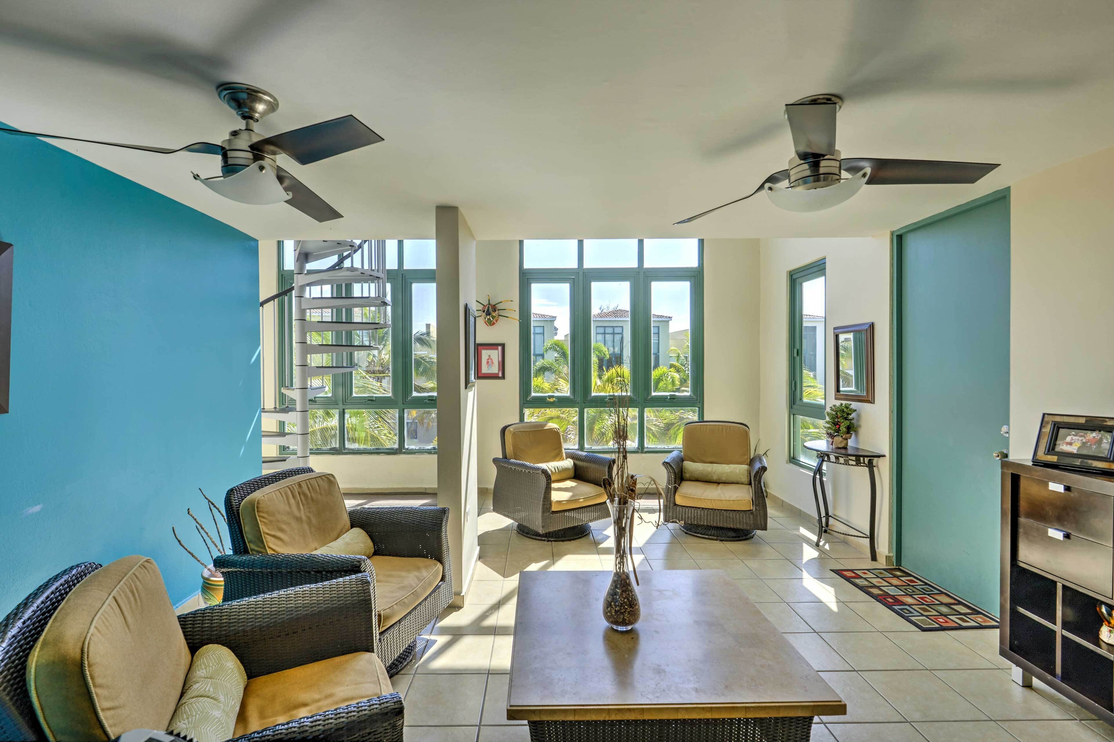 A spacious living area with wicker furniture, a coffee table, and ceiling fans. Large windows provide natural light and views of greenery outside.