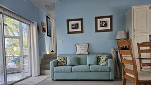 Een woonkamer met een lichtblauwe bank, twee ingelijste foto's aan de muur, een houten stoel en een trap naar een buitenruimte.