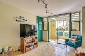 A living room with a television, a wooden entertainment unit, a teal armchair, and a sliding glass door leading to a balcony.