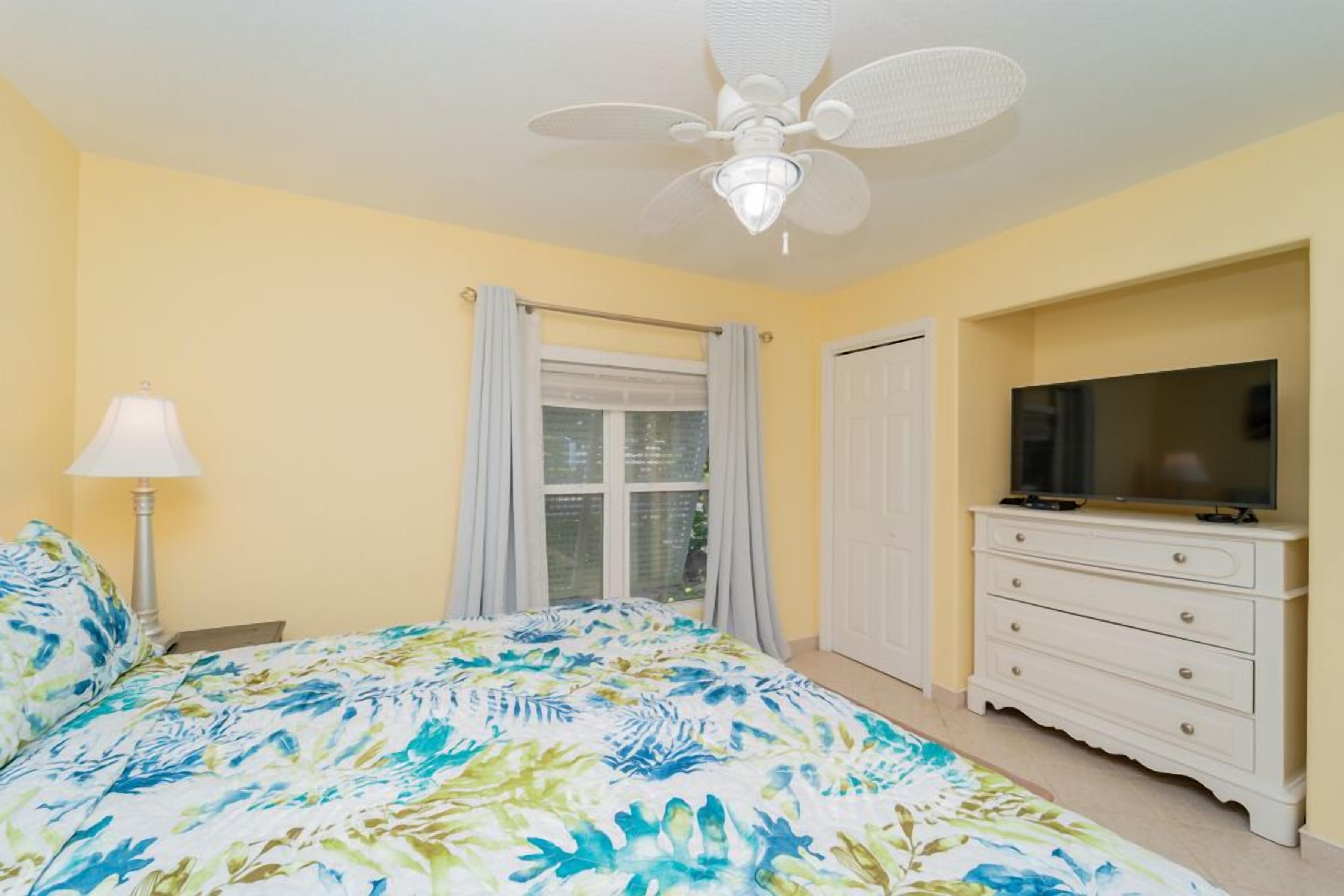A bedroom with a bed, a ceiling fan, a TV, and a window with curtains.