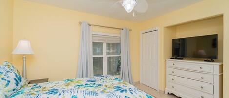 A bedroom with a bed, a ceiling fan, a TV, and a window with curtains.