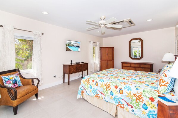 Une chambre à coucher comprenant un lit, une chaise, un bureau, une armoire, un ventilateur de plafond et une fenêtre avec des rideaux.