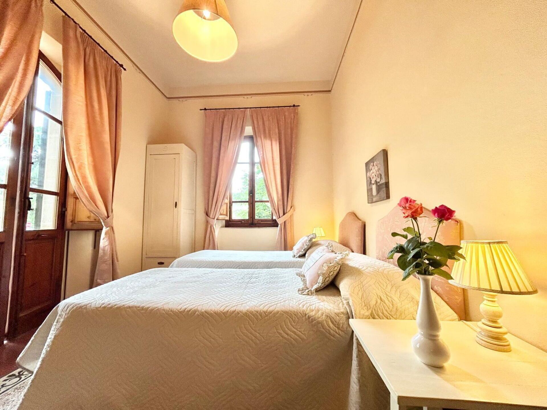 Quarto de hotel com duas camas, um guarda-roupa, uma janela com cortinas, uma mesinha de cabeceira com abajur e um vaso com flores.