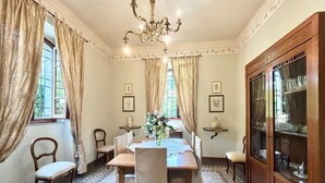 Une salle à manger élégante, agrémentée d’un lustre, de grandes fenêtres avec rideaux, d’un buffet en bois contenant de la verrerie, et d’un tapis à motifs.