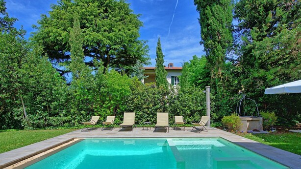 Une piscine rectangulaire, bordée d’une terrasse carrelée et entourée de verdure et d’arbres.