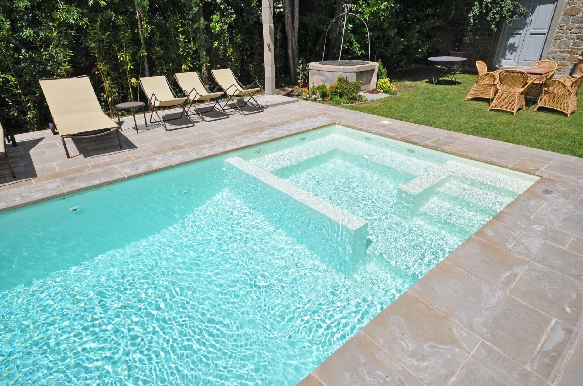 A rectangular swimming pool with clear water, surrounded by a stone deck. There are lounge chairs and a small table with chairs in the background.