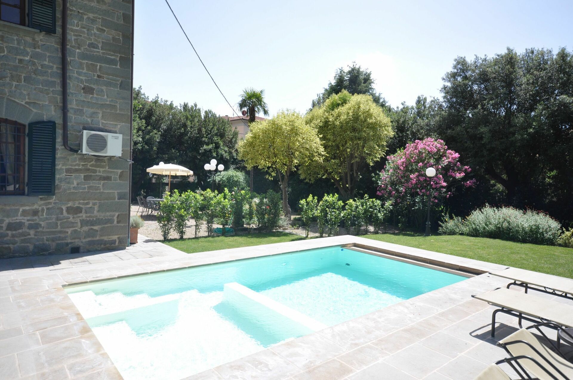 Una piscina con solárium de piedra, rodeada por un jardín con diversos árboles y arbustos.