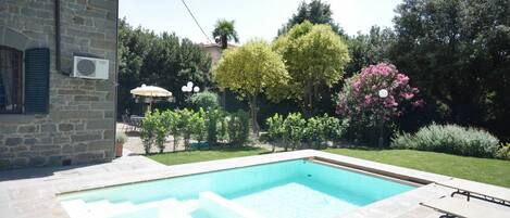 Una piscina con solárium de piedra, rodeada por un jardín con diversos árboles y arbustos.