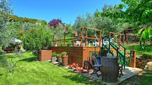 Casetta di legno rustica con uno scivolo e una rete da arrampicata, immersa nel verde e circondata da un muro in pietra.