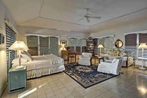 Une chambre d’hôtel avec deux lits, un bureau, une chaise, un canapé, un ventilateur de plafond et de grandes fenêtres équipées de stores.