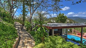 A stone pathway leading to a pool area with lush greenery and a mountain view. - Tropical Ponce Home: Private Pool & Outdoor Oasis! (Ponce)