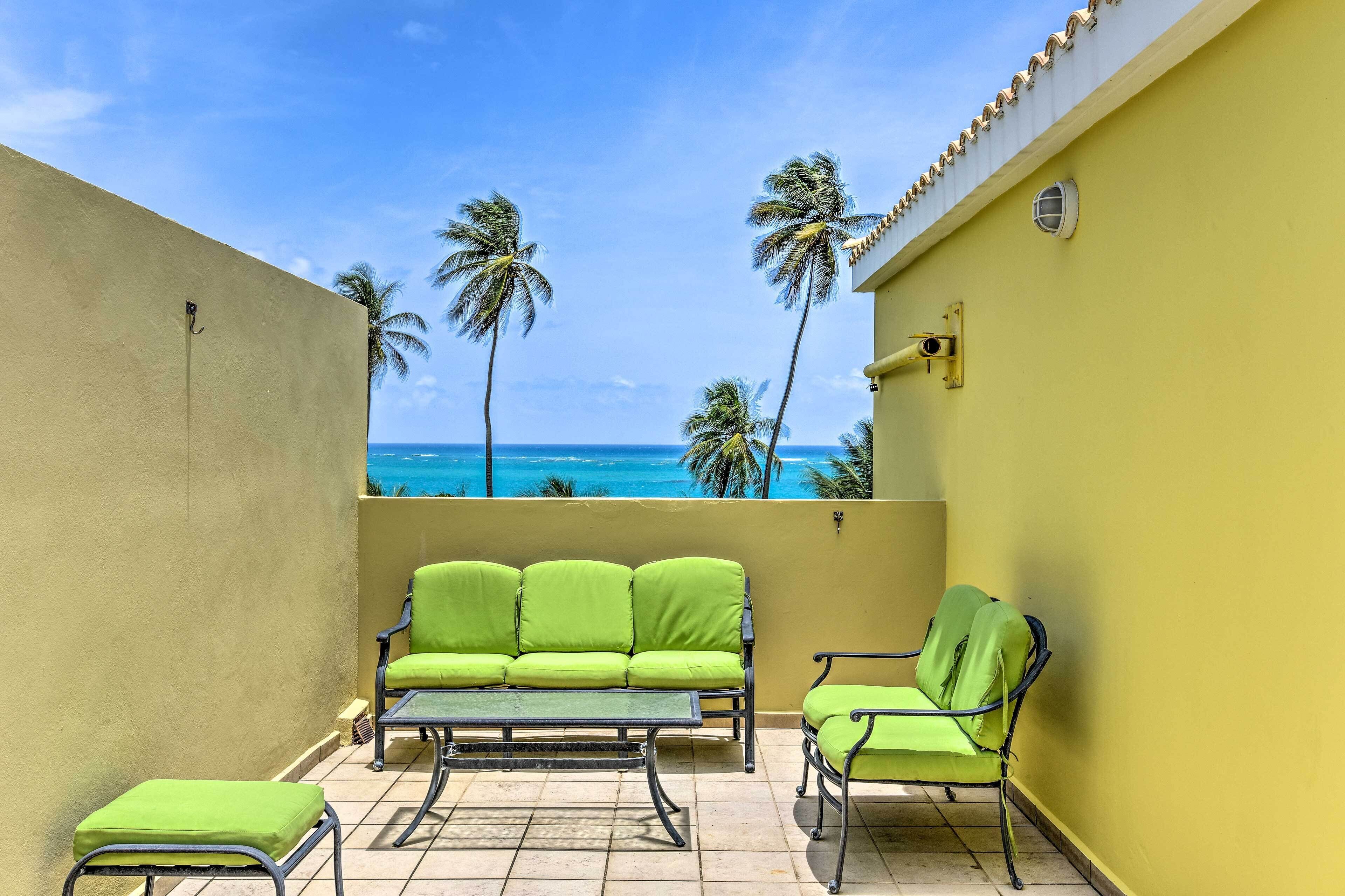 A balcony with a green sofa, a small table, and a chair, overlooking a beach with palm trees and the ocean.
