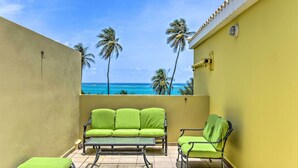Ein Balkon mit einem grünen Sofa, einem kleinen Tisch und einem Stuhl mit Blick auf einen Strand, Palmen und das Meer.