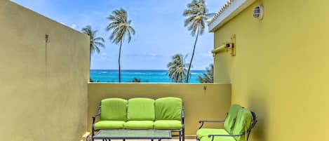 A balcony with a green sofa, a small table, and a chair, overlooking a beach with palm trees and the ocean.