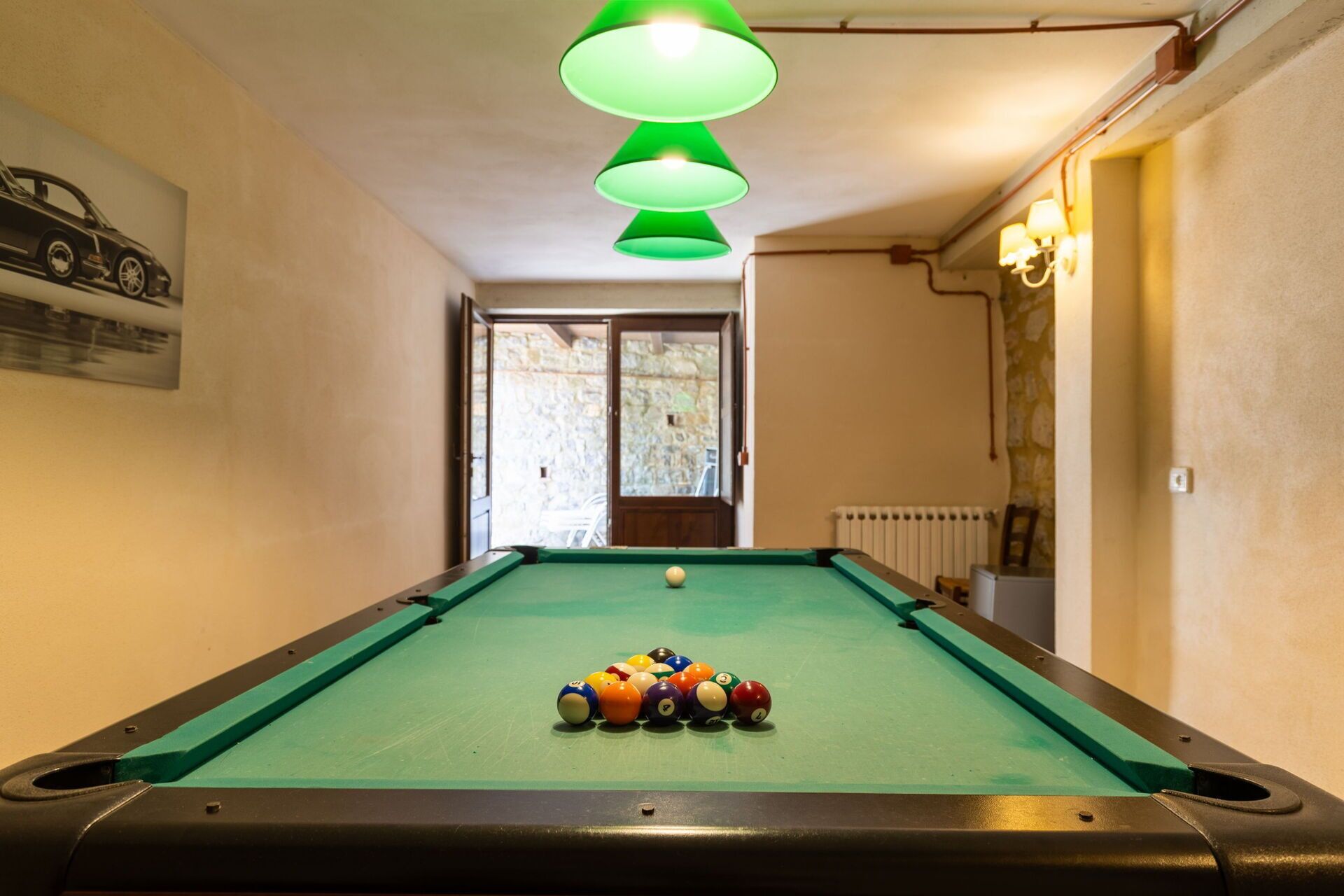 Une table de billard avec des billes, une lampe verte suspendue au plafond, une porte donnant sur l’extérieur et un radiateur au mur.