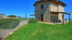 Casa de piedra con patio y parrilla exterior, rodeada por una cerca metálica y follaje.