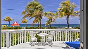 A balcony with white chairs and a table, overlooking a beach with palm trees and a red gazebo.