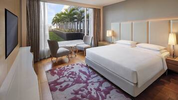 Habitación de hotel con una cama grande, un escritorio, una silla, un televisor y vistas a palmeras desde el exterior.