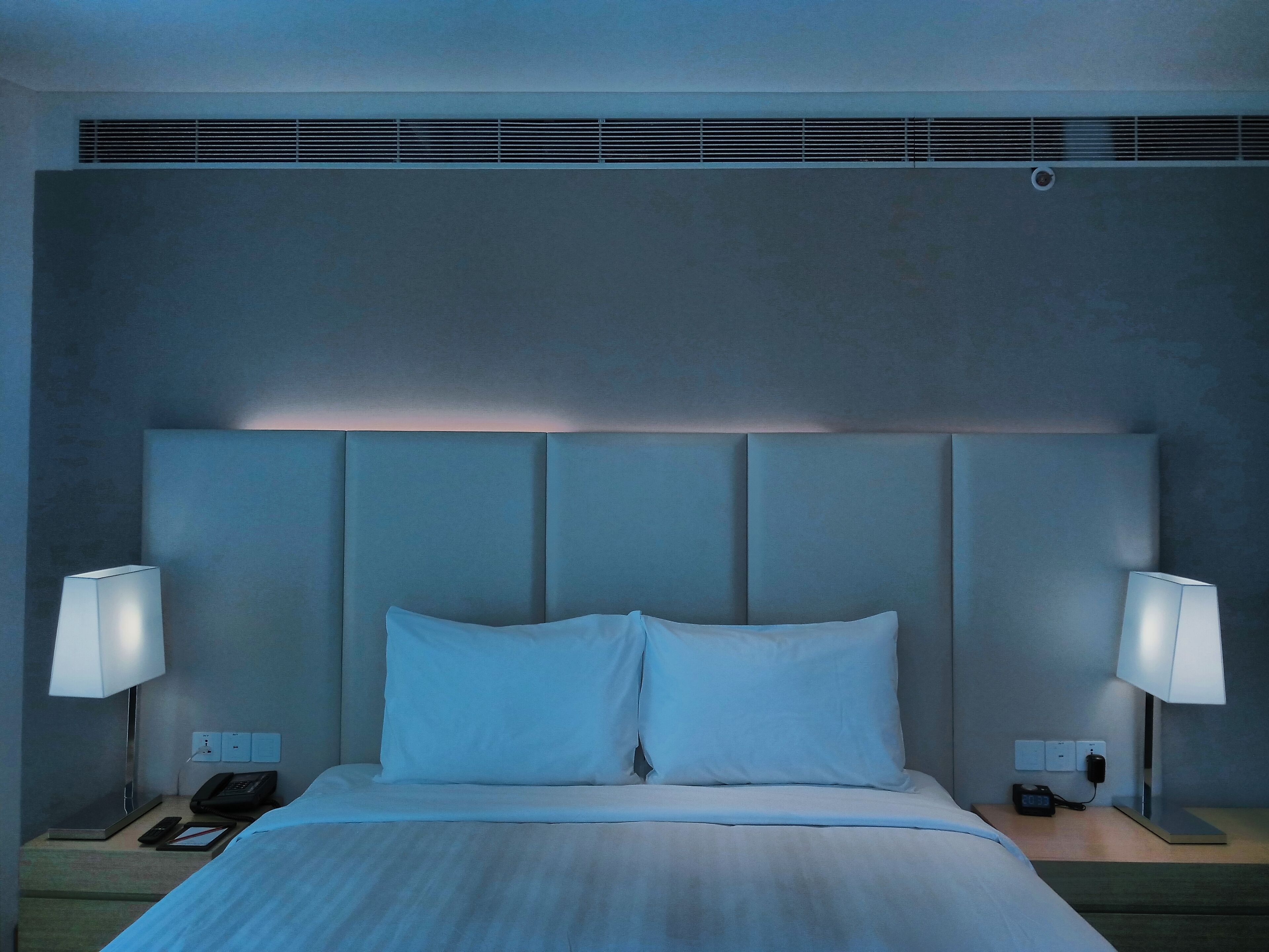 A hotel room with a large bed, two bedside lamps, a nightstand with a phone and a book, and a wall-mounted vent.