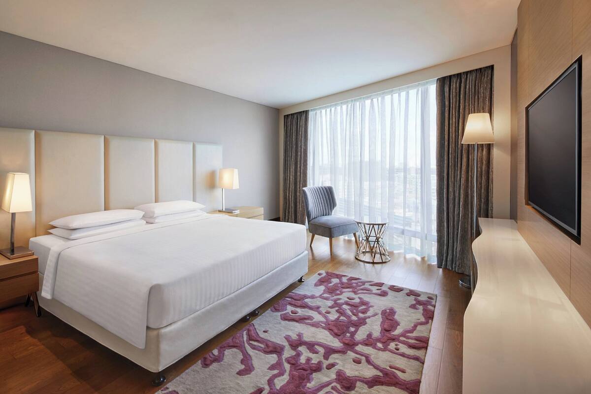 Ein modernes Hotelzimmer mit einem großen Bett, zwei Nachttischen mit Lampen, einem Sessel, einem Fernseher und einem großen Fenster mit Vorhängen.