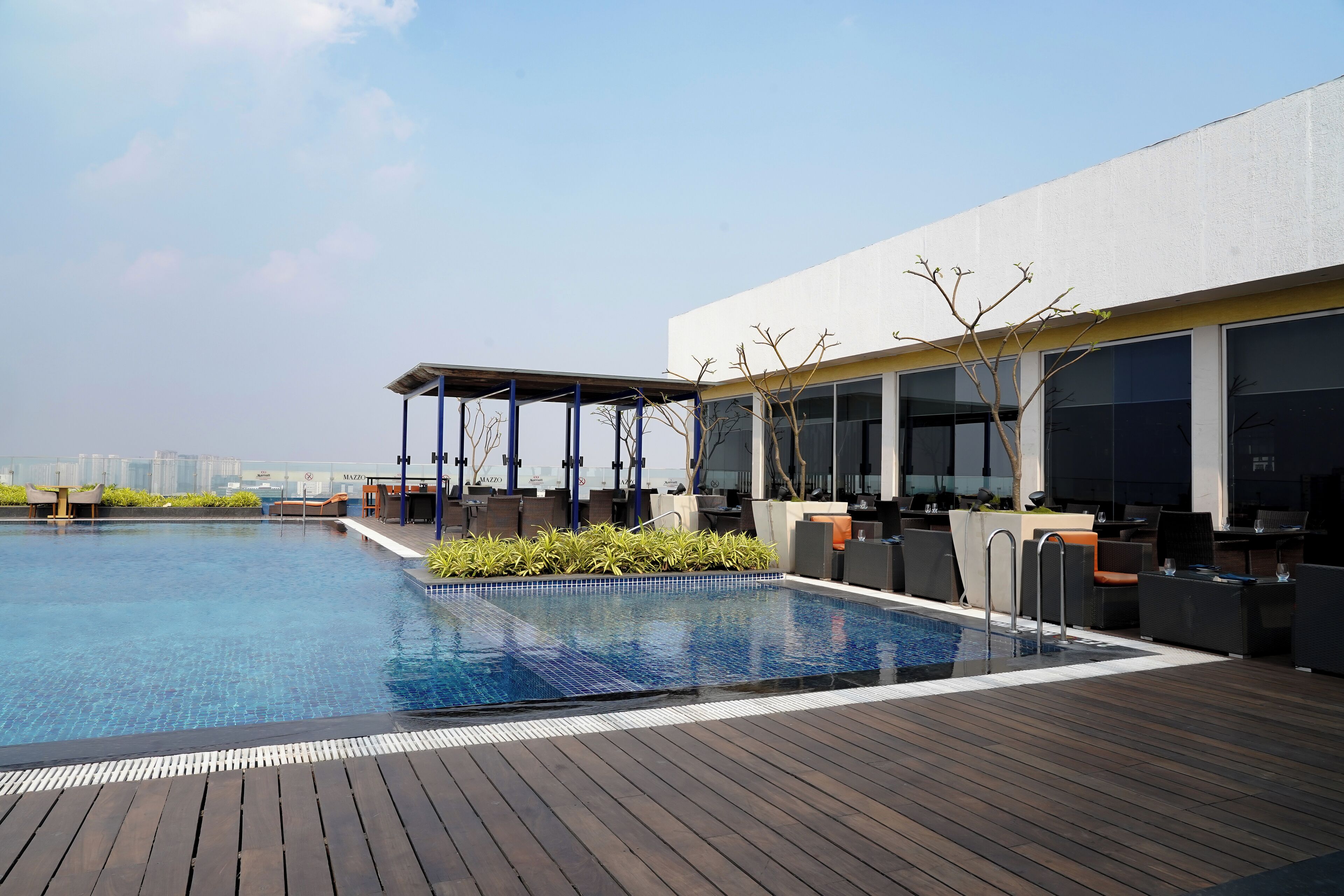 A rooftop swimming pool with wooden decking, outdoor seating, and a covered area with a view of the city skyline.