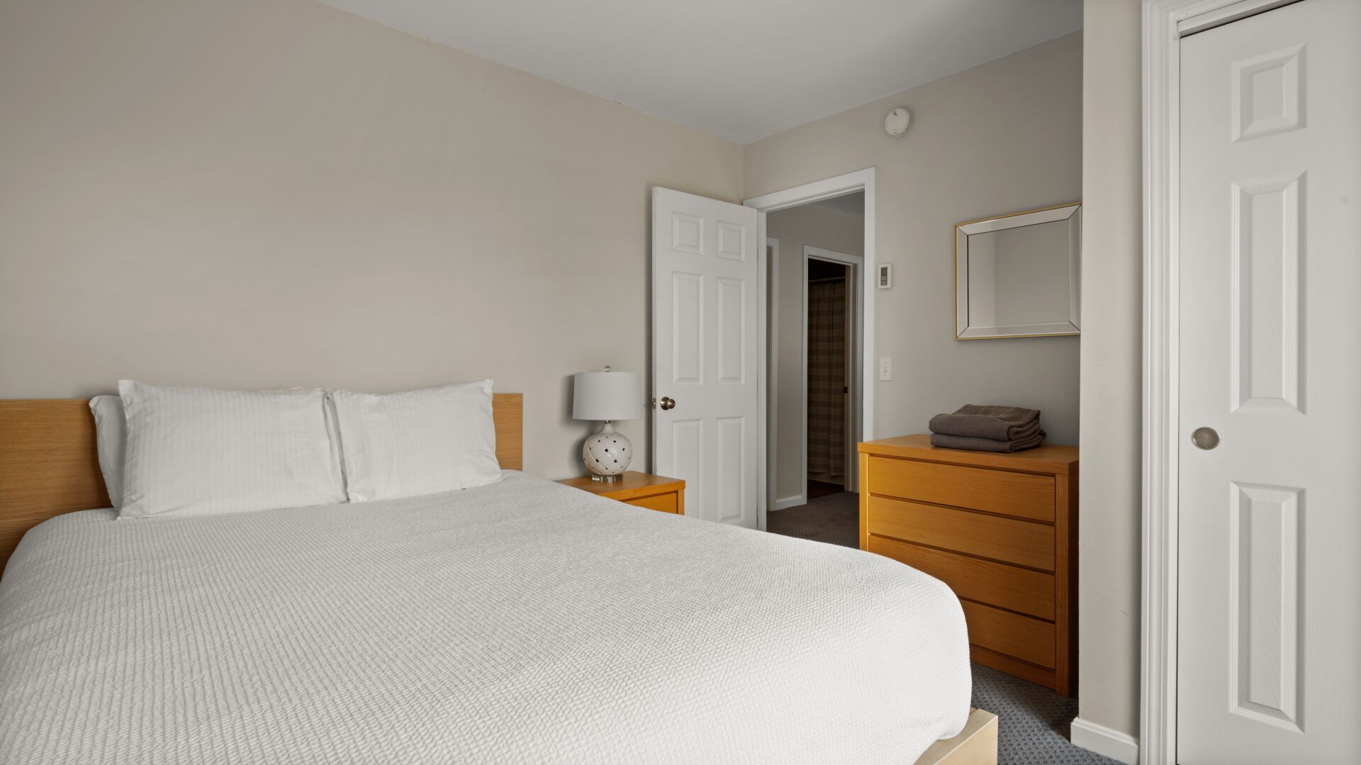 A neatly arranged bedroom with a large bed, a bedside table with a lamp, a wooden dresser, and a door leading to a closet.
