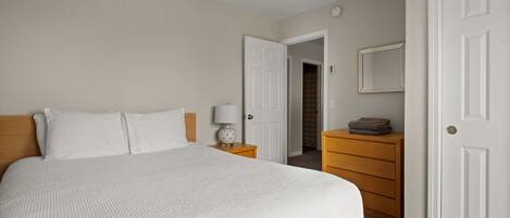 A neatly arranged bedroom with a large bed, a bedside table with a lamp, a wooden dresser, and a door leading to a closet.