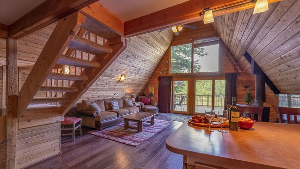 Ein gemütliches Blockhaus-Interieur mit einer Holztreppe, einer Sofagarnitur, einem Essbereich und einem großen Fenster mit Blick ins Freie.
