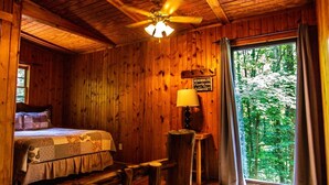 A wooden cabin room with a bed, a ceiling fan, a window with curtains, and a sign on the wall.