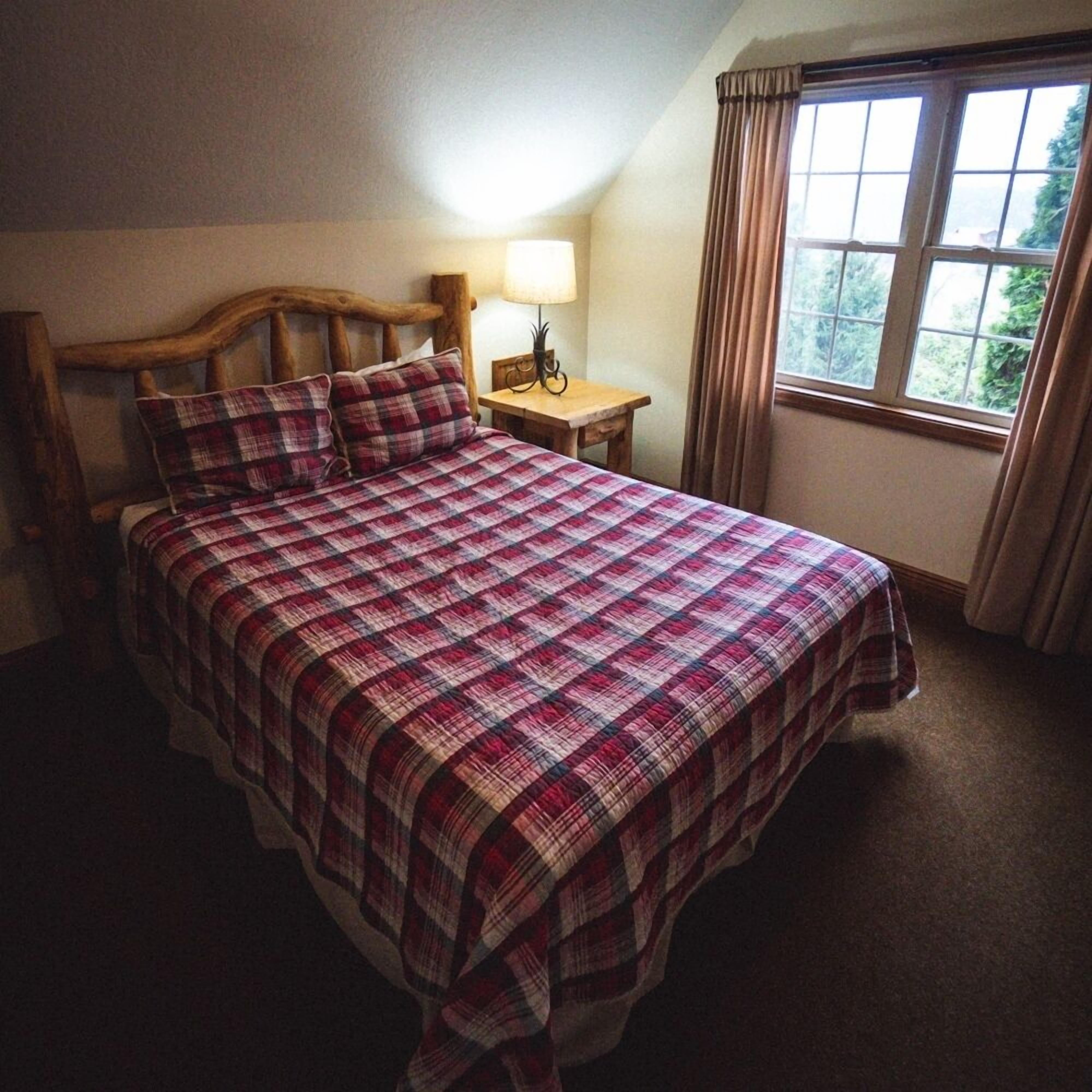 Una cama bien hecha con colcha escocesa, cabecera de madera, mesita de noche con lámpara y ventana con cortinas.