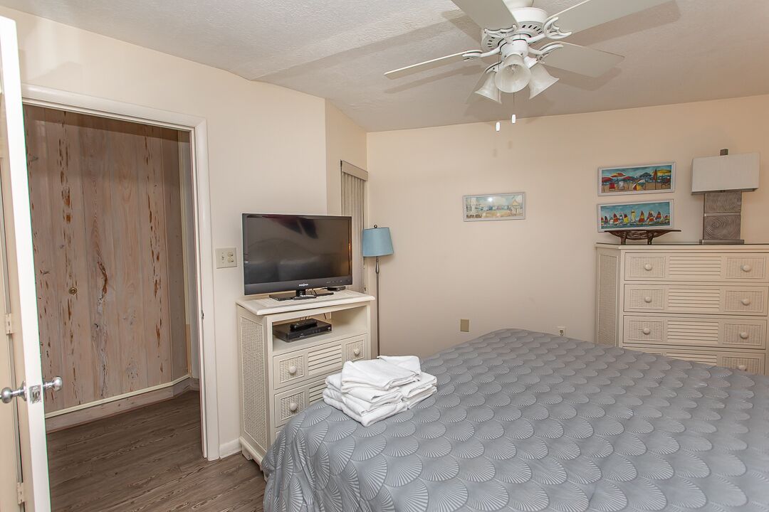 A bedroom with a bed, a television on a stand, a ceiling fan, a dresser, and a door.
