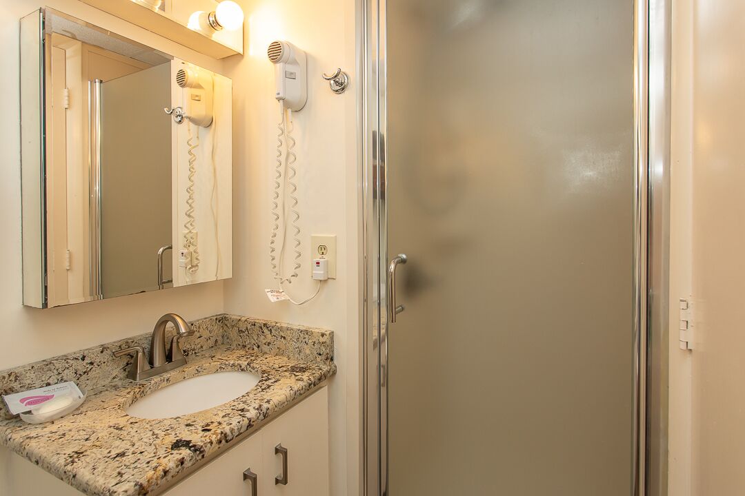 A bathroom with a granite countertop sink, a mirror, a hairdryer, and a glass shower enclosure.
